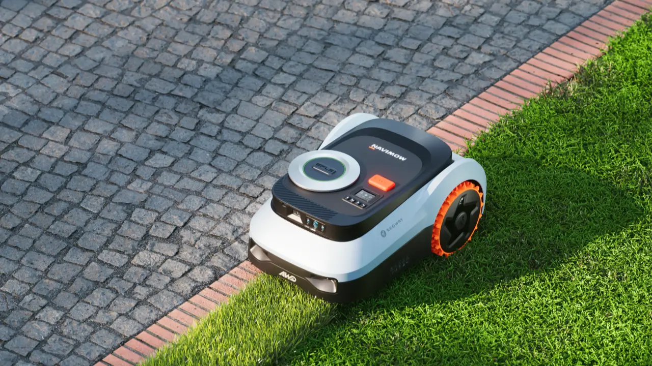 De Navimow robotmaaier met oranje wielen en een gladde witte behuizing, die automatisch het gras aan de rand van het gazon maait waar het overgaat in de kasseistrook.